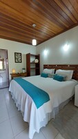 Un dormitorio con techo de madera, cama con ropa de cama blanca y verde azulado, mesita de noche, luz fijada en la pared y armario de madera.