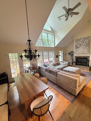 A spacious living area with a high ceiling, a fireplace, a large window, and a dining area with a wooden table and chairs. - Ski's The Day: Winsome 3-bedroom mountain townhome with AC in Jiminy Peak (Hancock)