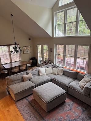 A spacious living room with a large sectional sofa, a dining table with chairs, a chandelier, and large windows with a view of trees. - Ski's The Day: Winsome 3-bedroom mountain townhome with AC in Jiminy Peak (Hancock)