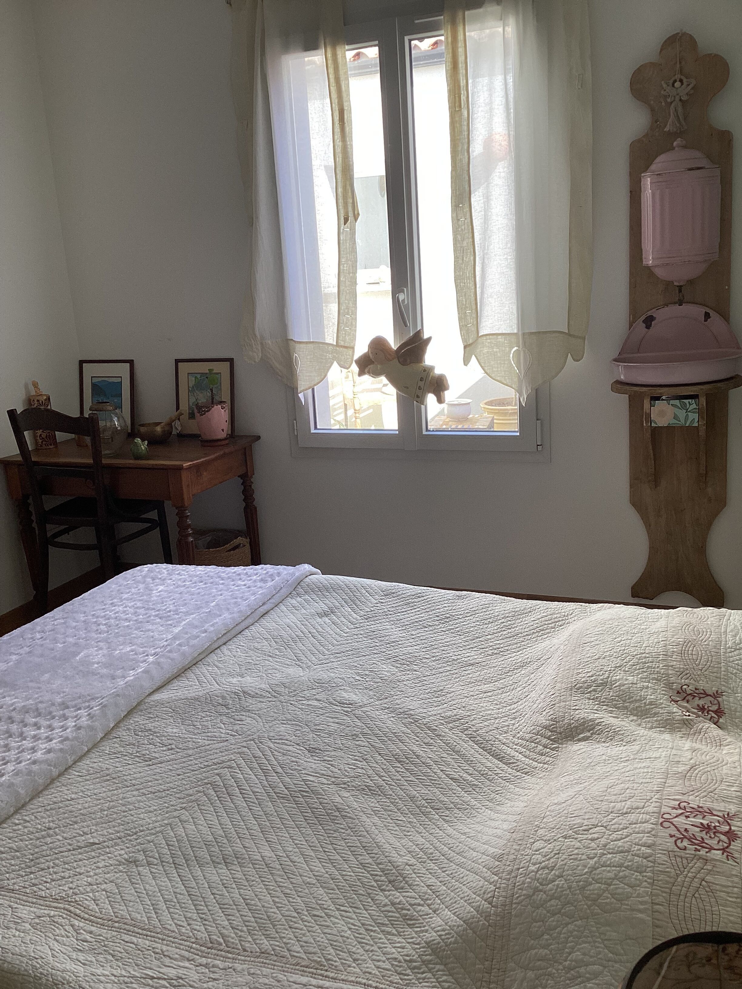 A bedroom with a bed, a wooden desk, a chair, a window with curtains, and a wall-mounted shelf with a pink container.