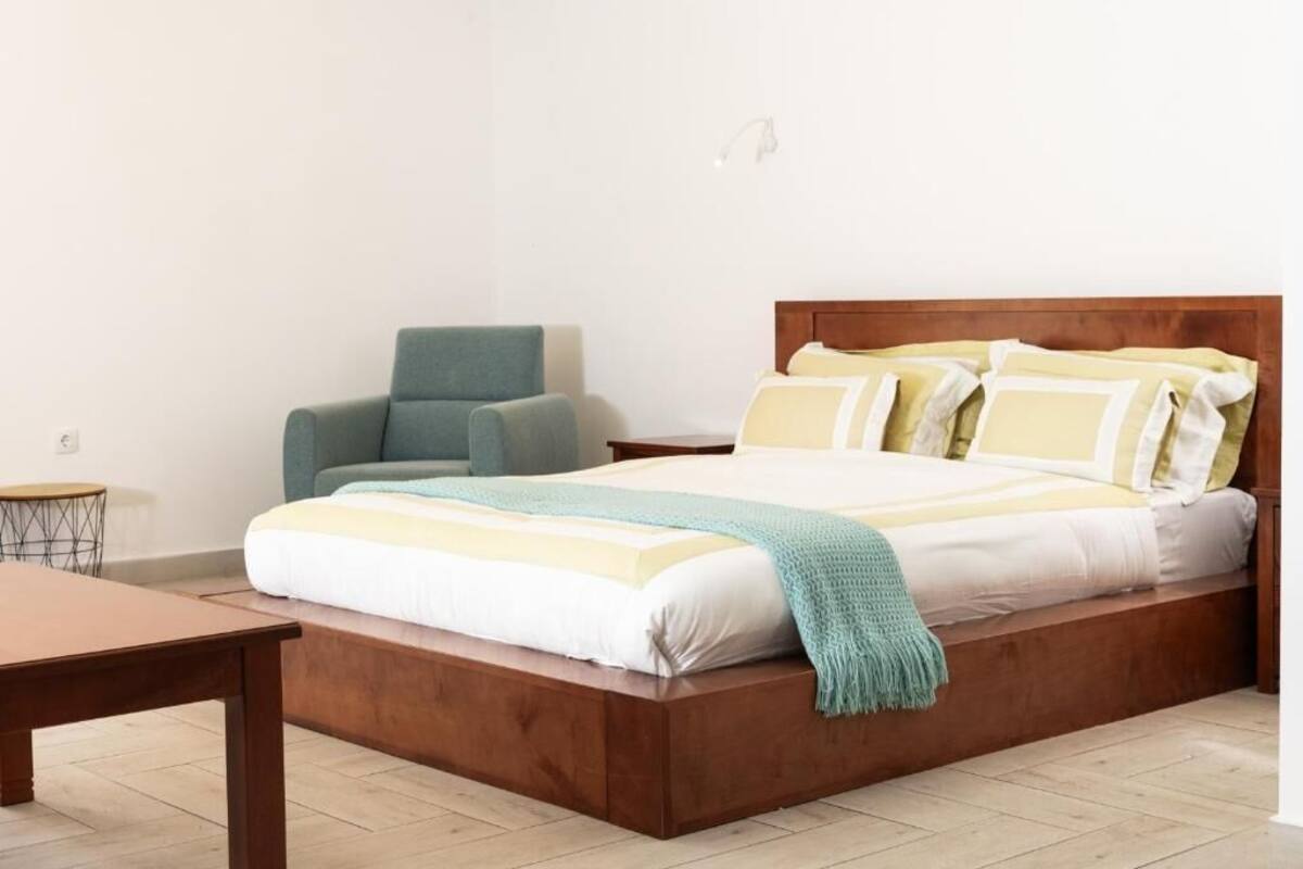 A single bed with a wooden headboard, a small round table, and a chair in a simple room with white walls and a tiled floor.