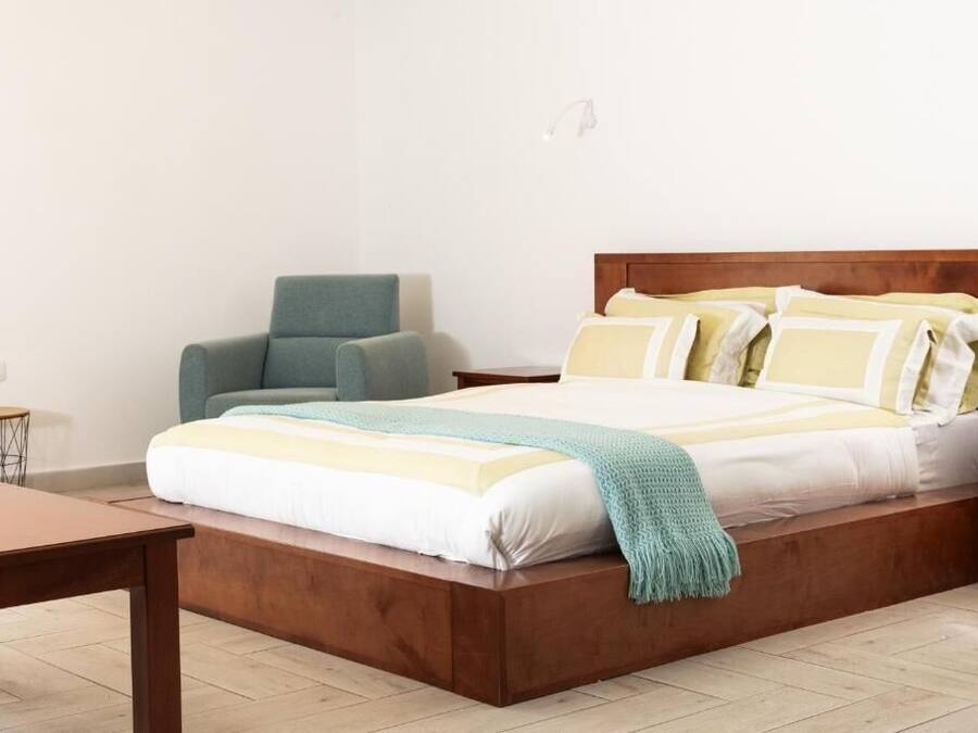 A single bed with a wooden headboard, a small round table, and a chair in a simple room with white walls and a tiled floor.
