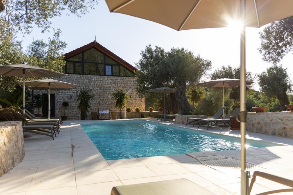 A swimming pool with lounge chairs and umbrellas, a stone building with a red roof, and trees in the background.