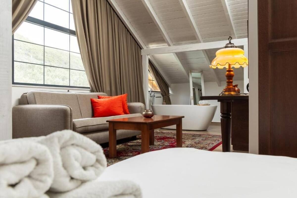 A hotel room with a sofa, a small table, a lamp, a bathtub, and a large window.