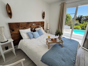Ein Schlafzimmer mit hölzernem Kopfteil, weißer Bettwäsche, einem Nachttisch mit Lampe und einem Tablett mit Frühstück, mit Blick auf einen Swimmingpool.