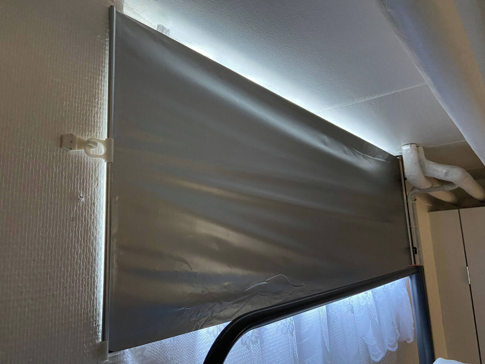 A room with a window covered by a grey blind, a white wall, and a white ceiling with a visible pipe.