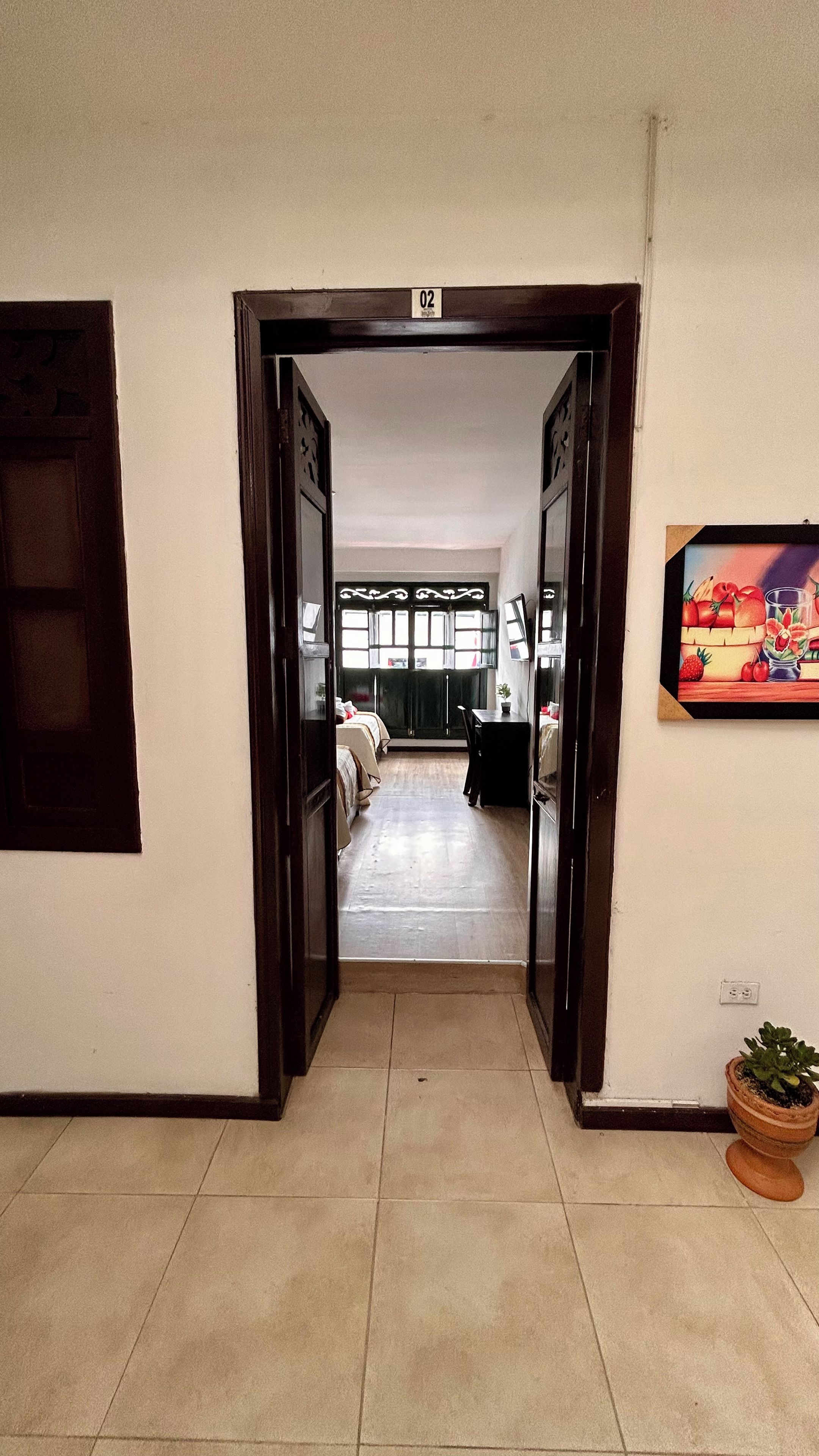 A hallway leading to a room with a bed, a desk, and a window. There is a door labeled '02' and a wall-mounted picture.