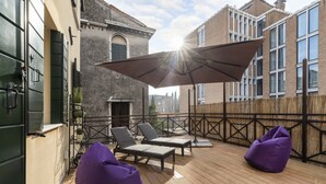 Un terrazzo in legno con poltrone relax e un pouf viola, un ombrellone grande e un edificio con facciata in mattoni sullo sfondo.