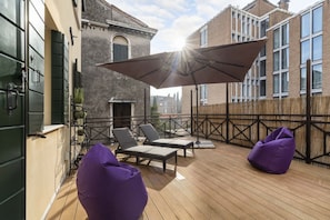 Eine Holzterrasse mit Liegestühlen und einem violetten Sitzsack, einem großen Sonnenschirm und einem Gebäude mit Backsteinfassade im Hintergrund.