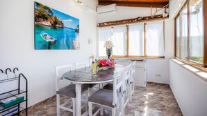 A dining area with a table and chairs, a painting of a coastal scene, and a window with a view. - Villa Luna with Pool and Mountain Views (Alcúdia)