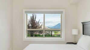 Ein Schlafzimmer mit großem Fenster, einem Bett, einem Nachttisch und einer Lampe.