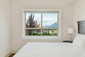 Ein Schlafzimmer mit großem Fenster, einem Bett, einem Nachttisch und einer Lampe.