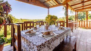 A covered outdoor dining area with a table set for four, surrounded by wooden chairs, with a view of a lush landscape.