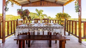A covered outdoor dining area with a table set for four, surrounded by wooden railings and palm trees.