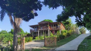 A two-story wooden house with a balcony, surrounded by lush greenery and a paved path.