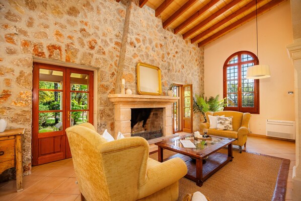 A stone-walled room with a fireplace, a sofa, a coffee table, and a window with a view of greenery.