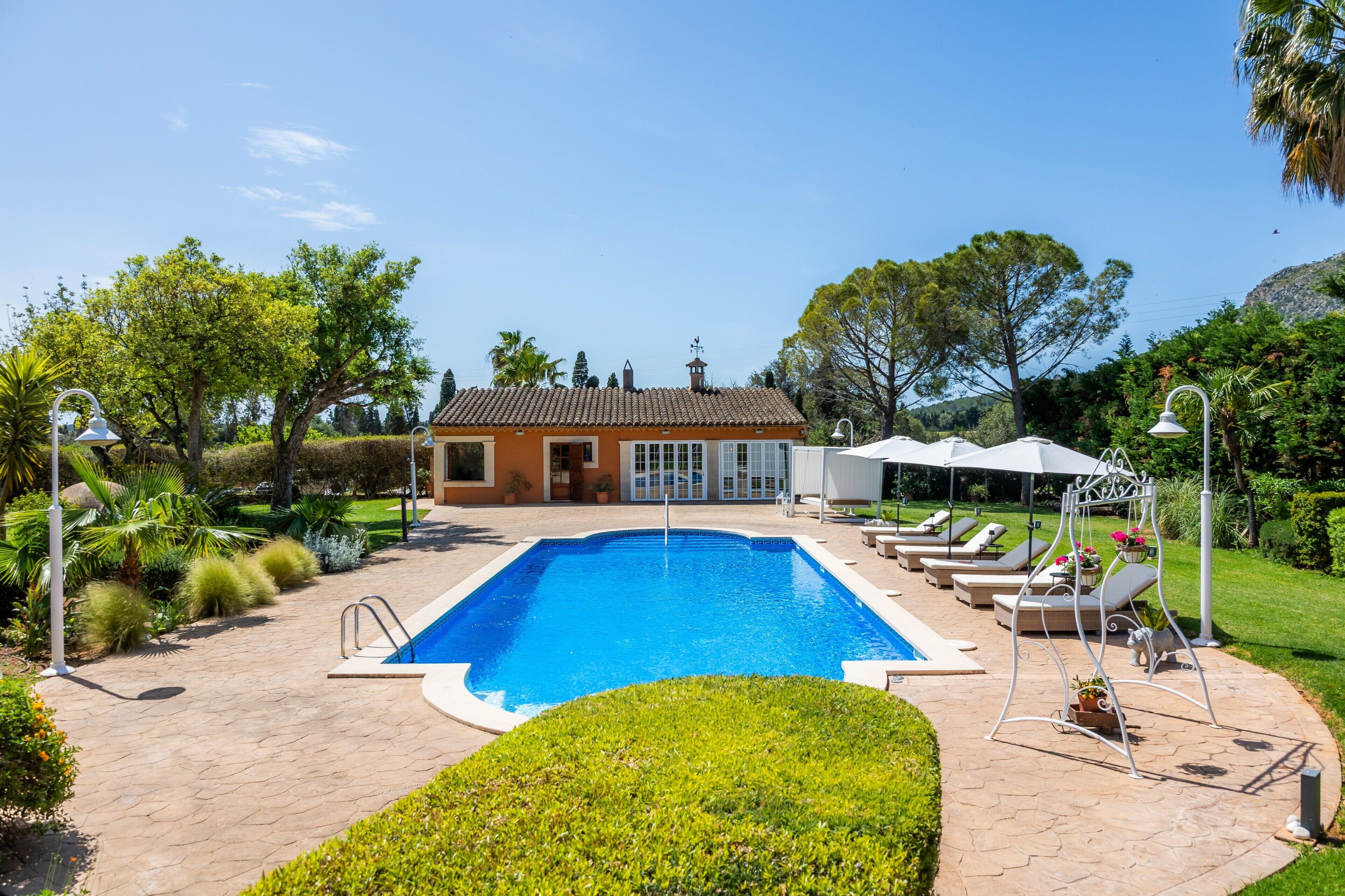 Una piscina rectangular con superficie de agua azul cristalina, rodeada por un patio empedrado con sillones de relax y sombrillas.