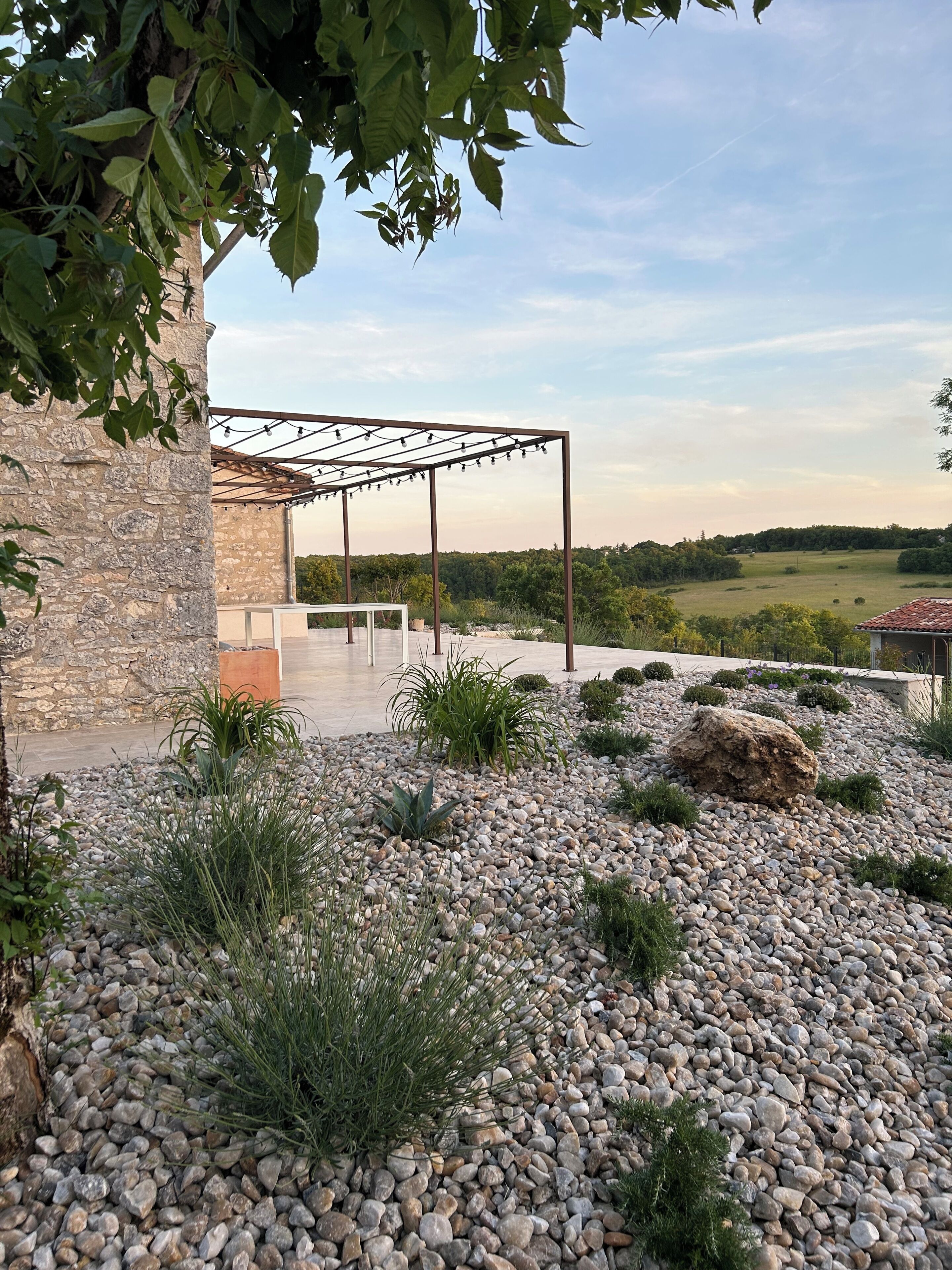 A stone building with a pergola, surrounded by a gravel garden with plants.