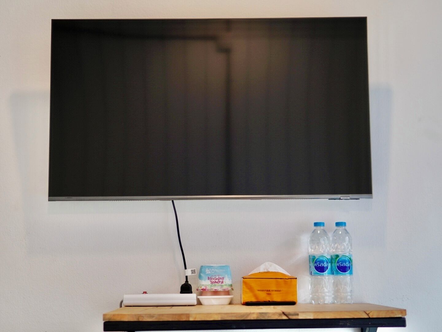 A flat-screen TV mounted on a wall, a wooden table with a tissue box, a water bottle, and a container of yogurt.
