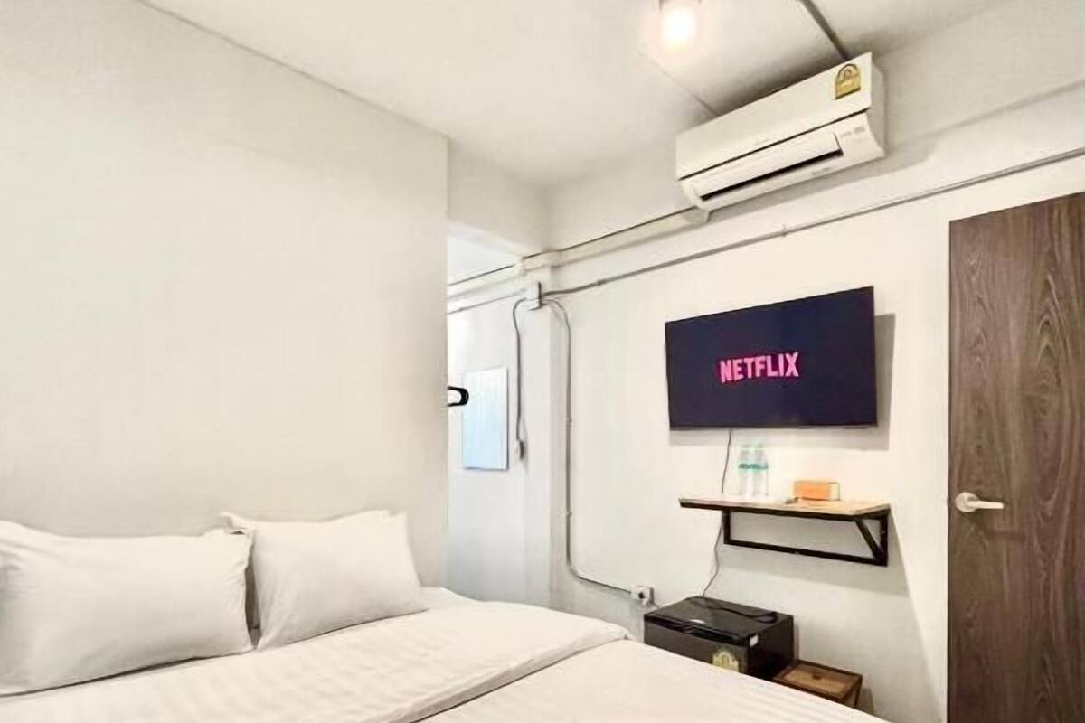 A modern hotel room with a large bed, a mounted television displaying 'NETFLIX,' a small table with a bottle, and an air conditioning unit.