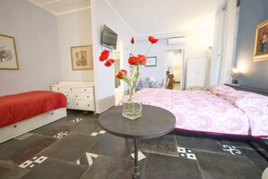 Ein Hotelzimmer mit einem Bett, einem kleinen Tisch mit einer Vase, einem Fernseher und einer Tür zu einem anderen Raum.