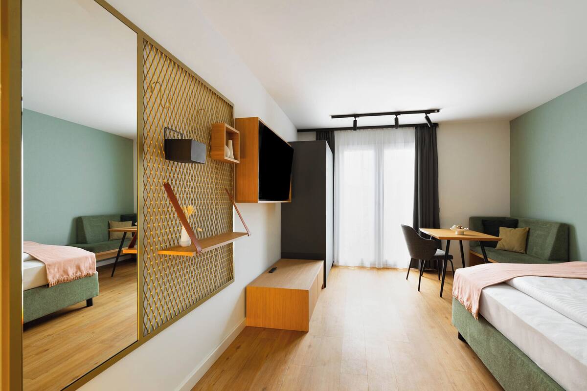Une chambre d’hôtel moderne avec un grand miroir, un plancher en bois, un canapé, un lit, un bureau et une télévision.