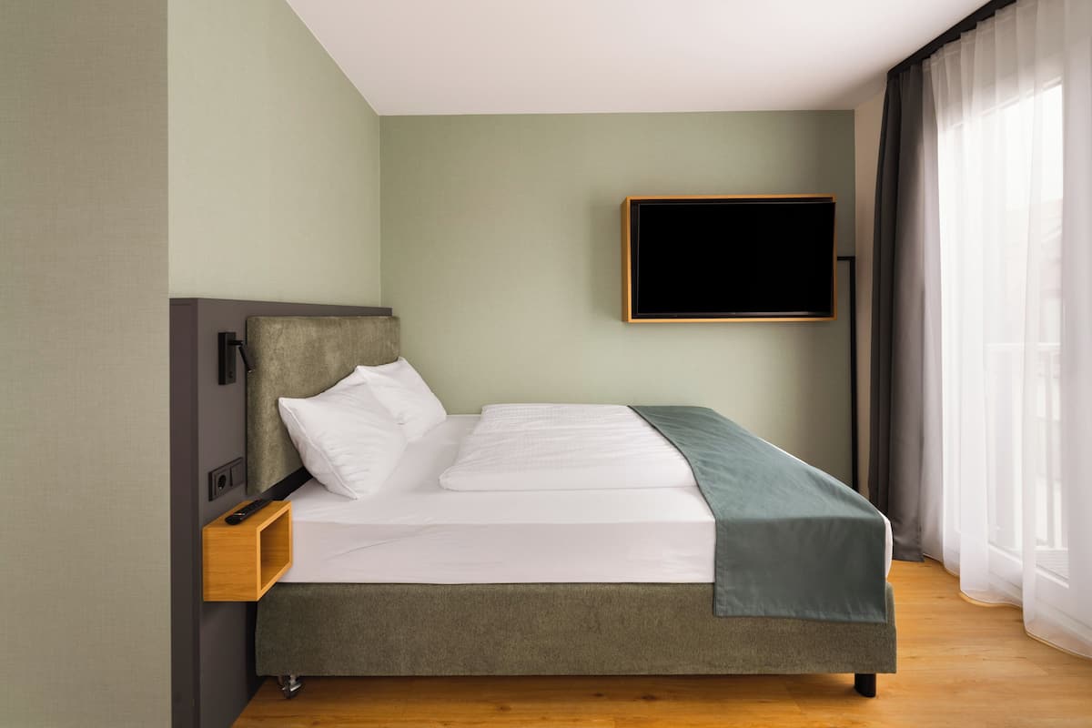 Un lit bien fait, avec une tête de lit grise, un téléviseur à écran plat fixé au mur et une table de chevet en bois sur laquelle se trouve une télécommande.