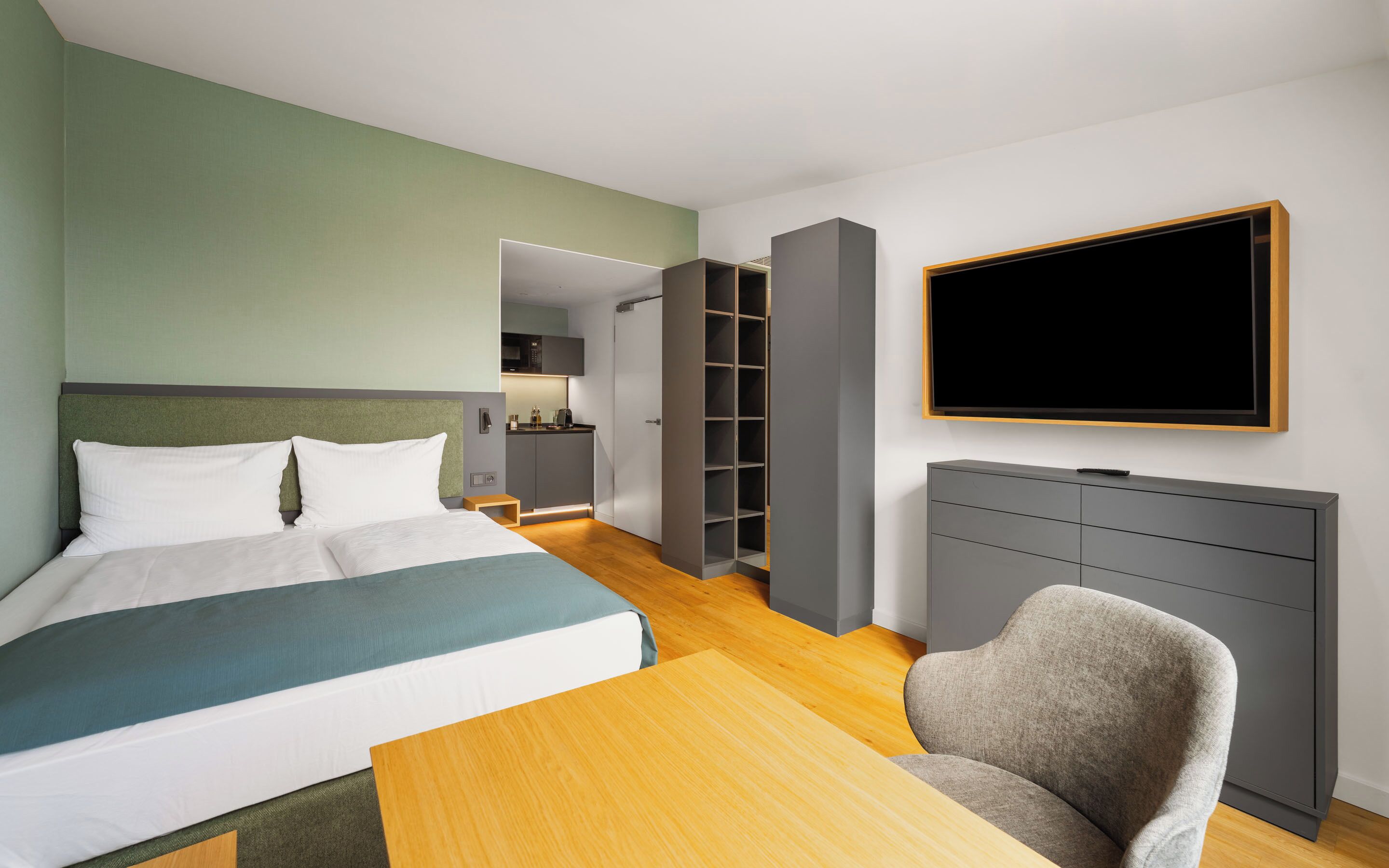 A hotel room with a bed, a desk, a chair, a TV, a wardrobe, and a kitchenette.