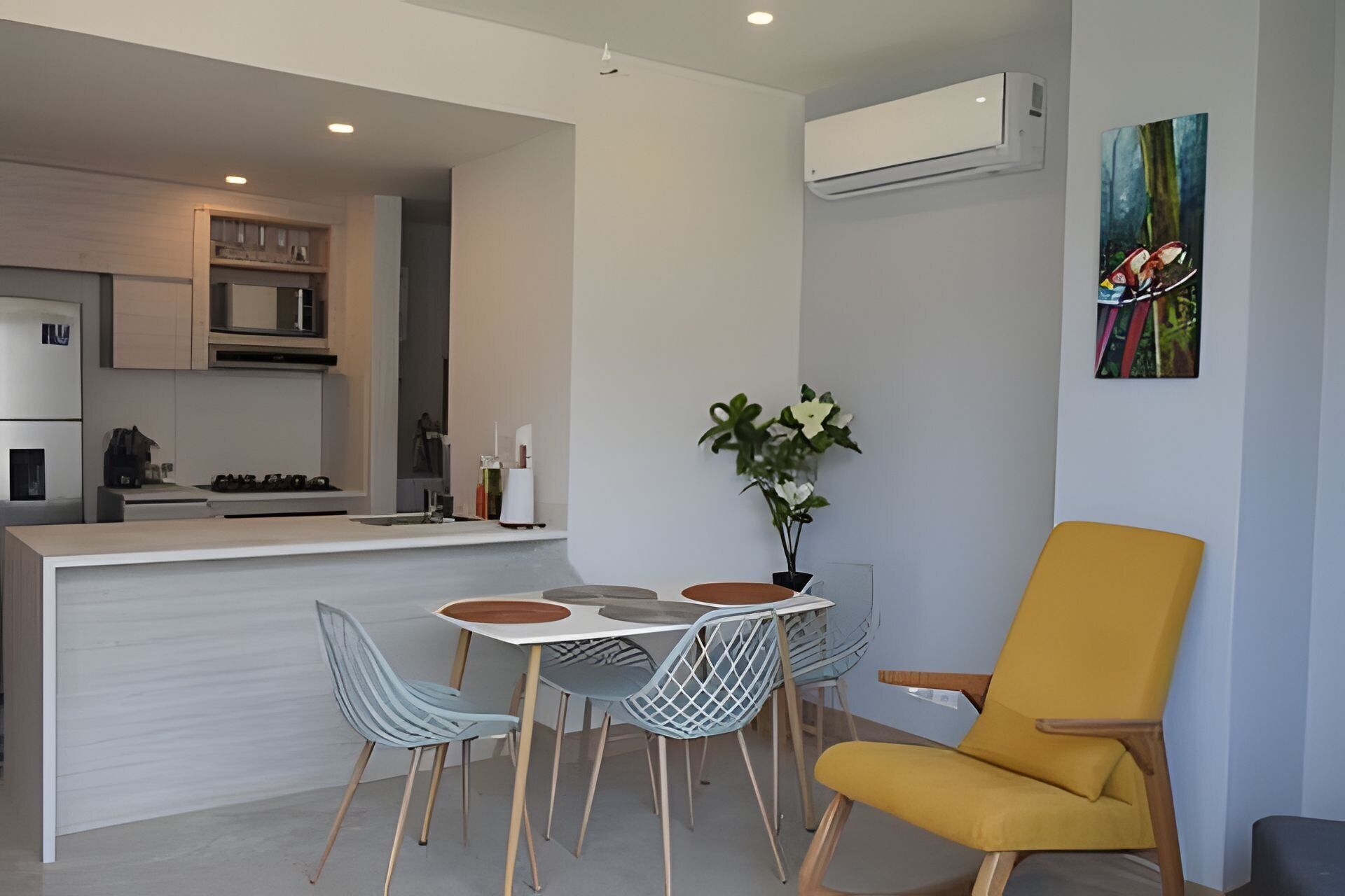 Une cuisine moderne avec des meubles blancs, un coin repas comprenant une table et des chaises, un fauteuil jaune et un appareil de climatisation.