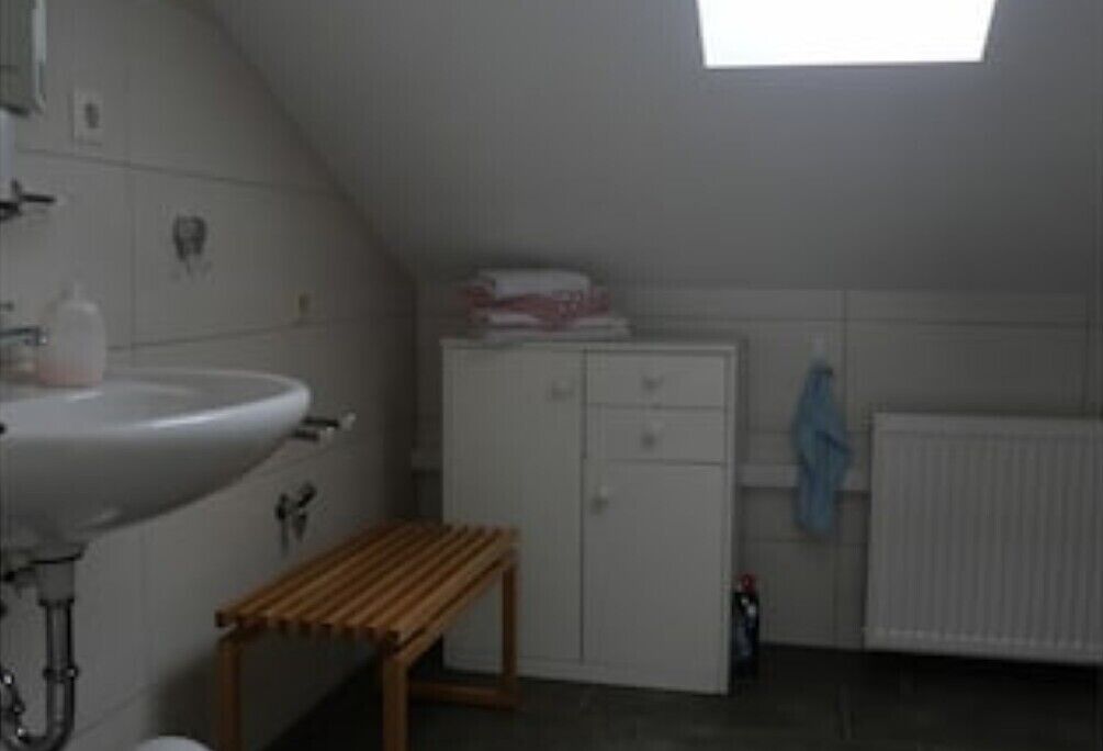 Ett litet, privat badrum med ett vitt handfat, en träbänk, ett skåp och ett takfönster.