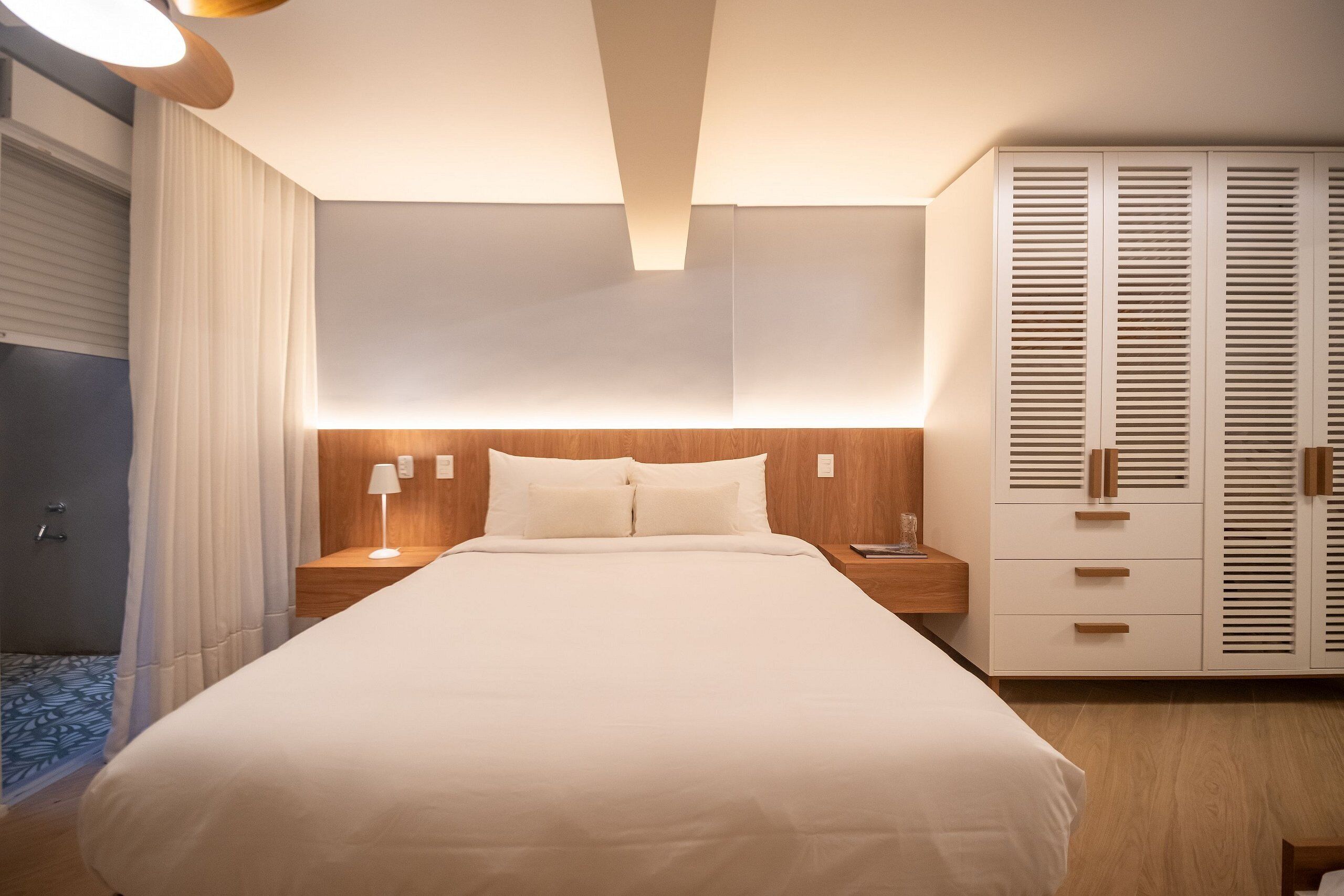Un lit bien fait, avec une tête de lit en bois, une lampe de chevet et une armoire aux lattes horizontales.