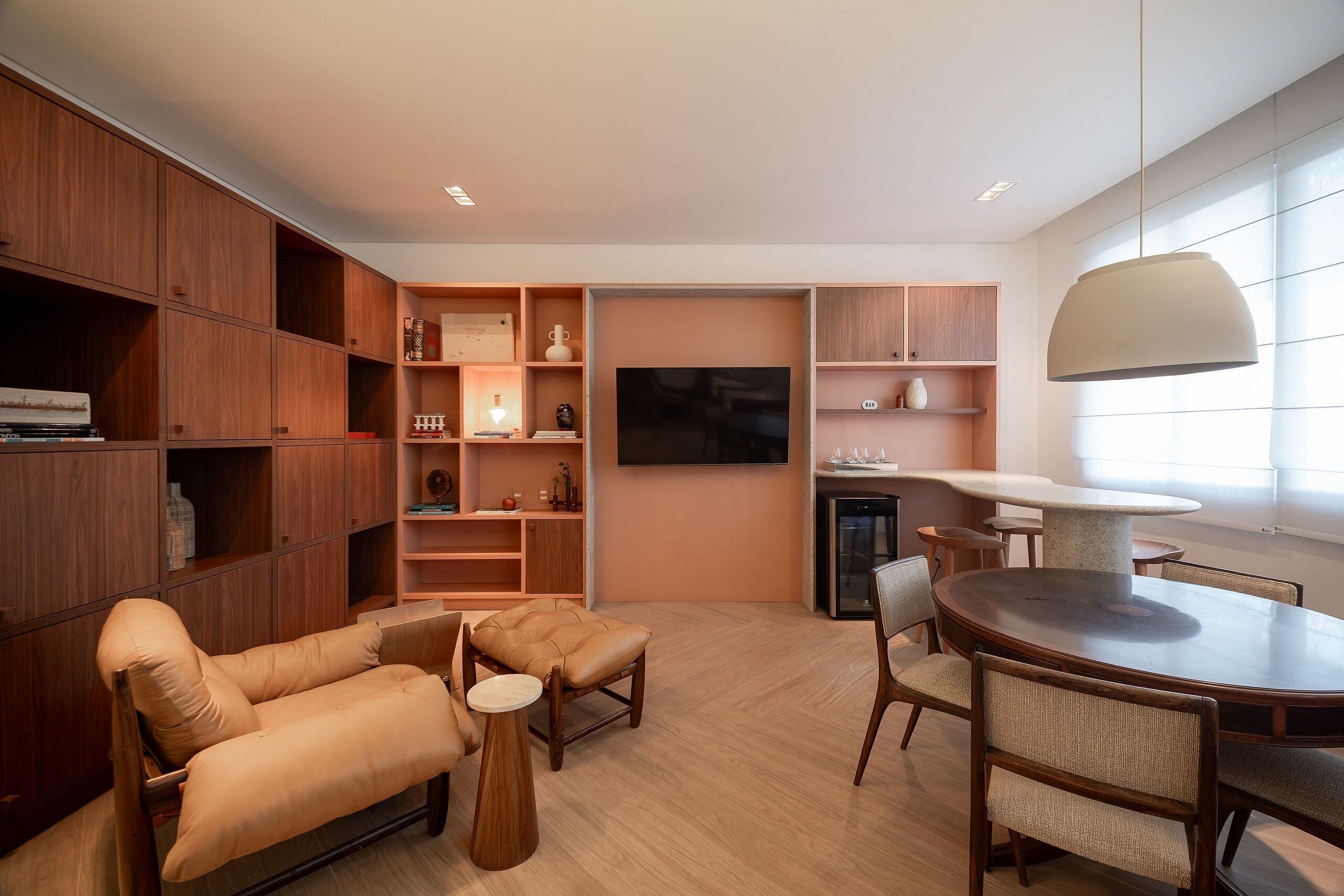 A modern living room with a sofa, ottoman, dining table, chairs, wooden shelving, and a flat-screen TV.