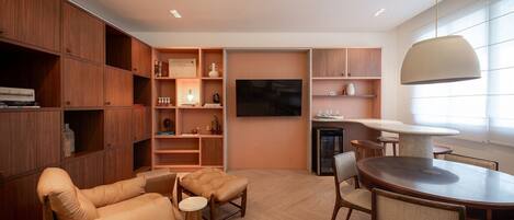 A modern living room with a sofa, ottoman, dining table, chairs, wooden shelving, and a flat-screen TV.