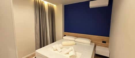 A modern hotel room with a bed, curtains, a blue accent wall, and an air conditioning unit.