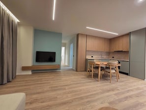 A modern living room with a flat-screen TV, a dining area with wooden chairs, and a kitchen with wooden cabinets. - Joanna Prestige Apartments (Sarande)