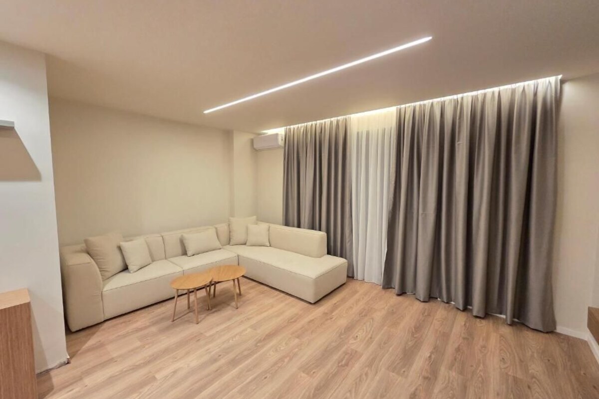 A modern living room with a sectional sofa, a wooden coffee table, and a large window with grey curtains.