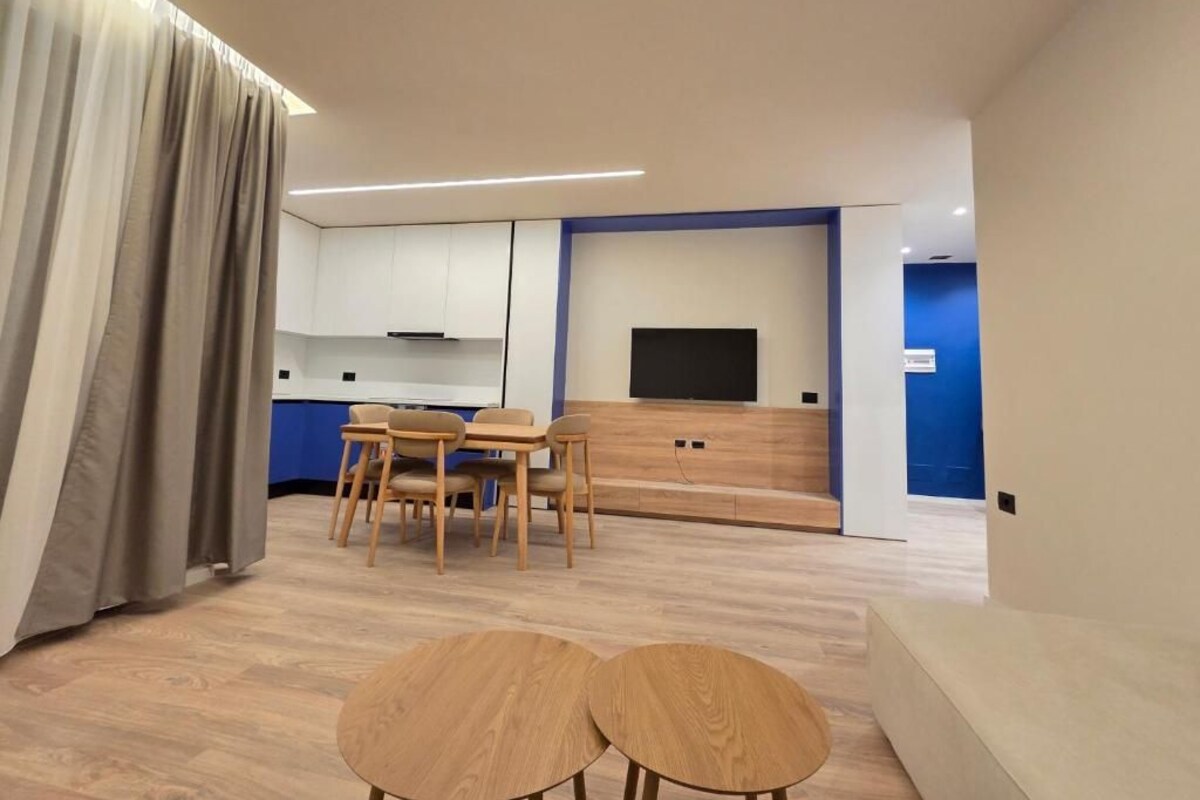 A modern living room with a sofa, two round tables, a dining area with chairs, a TV, and a kitchen in the background.