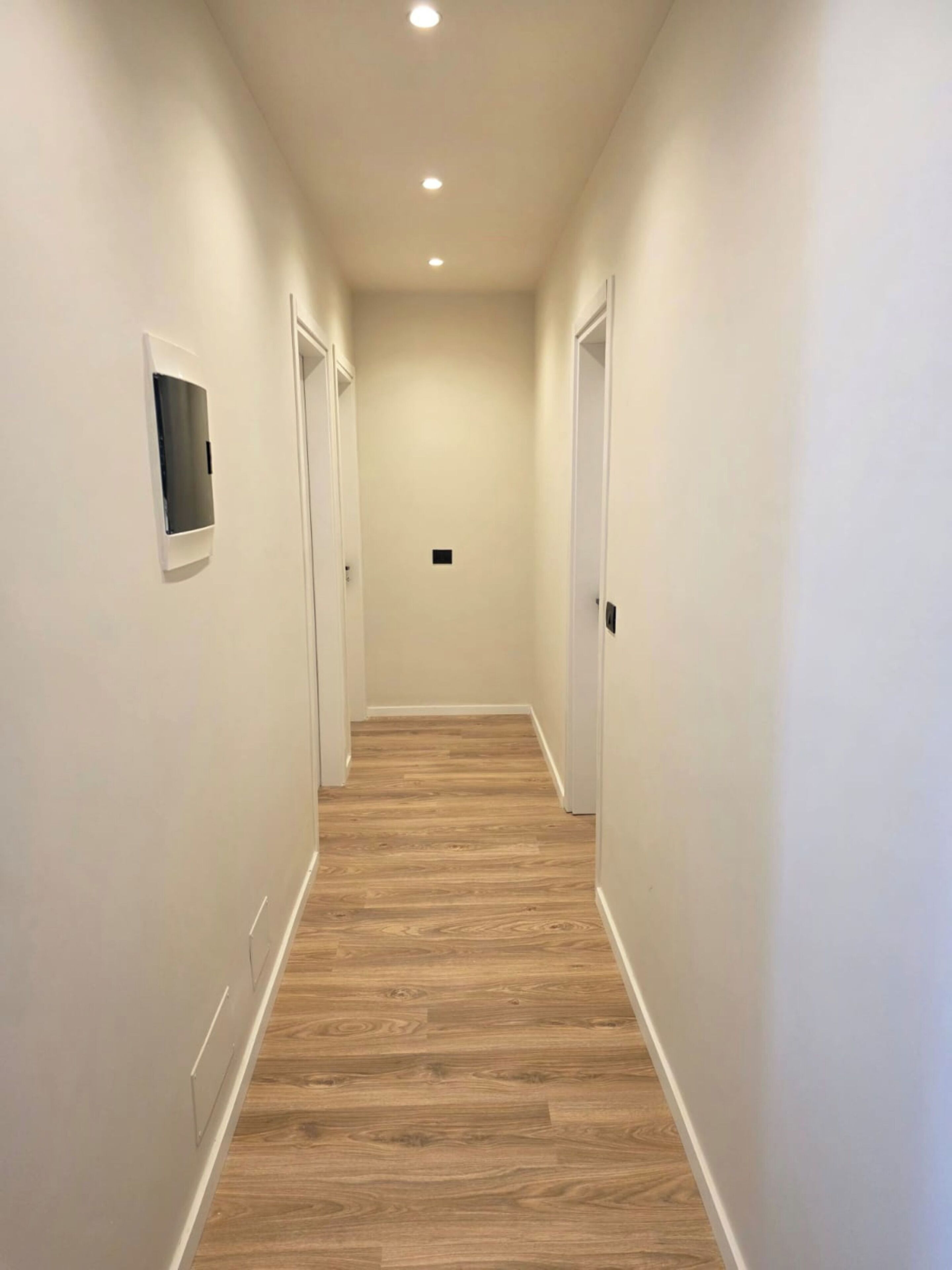 A narrow hallway with wooden flooring, white walls, and a recessed ceiling light.
