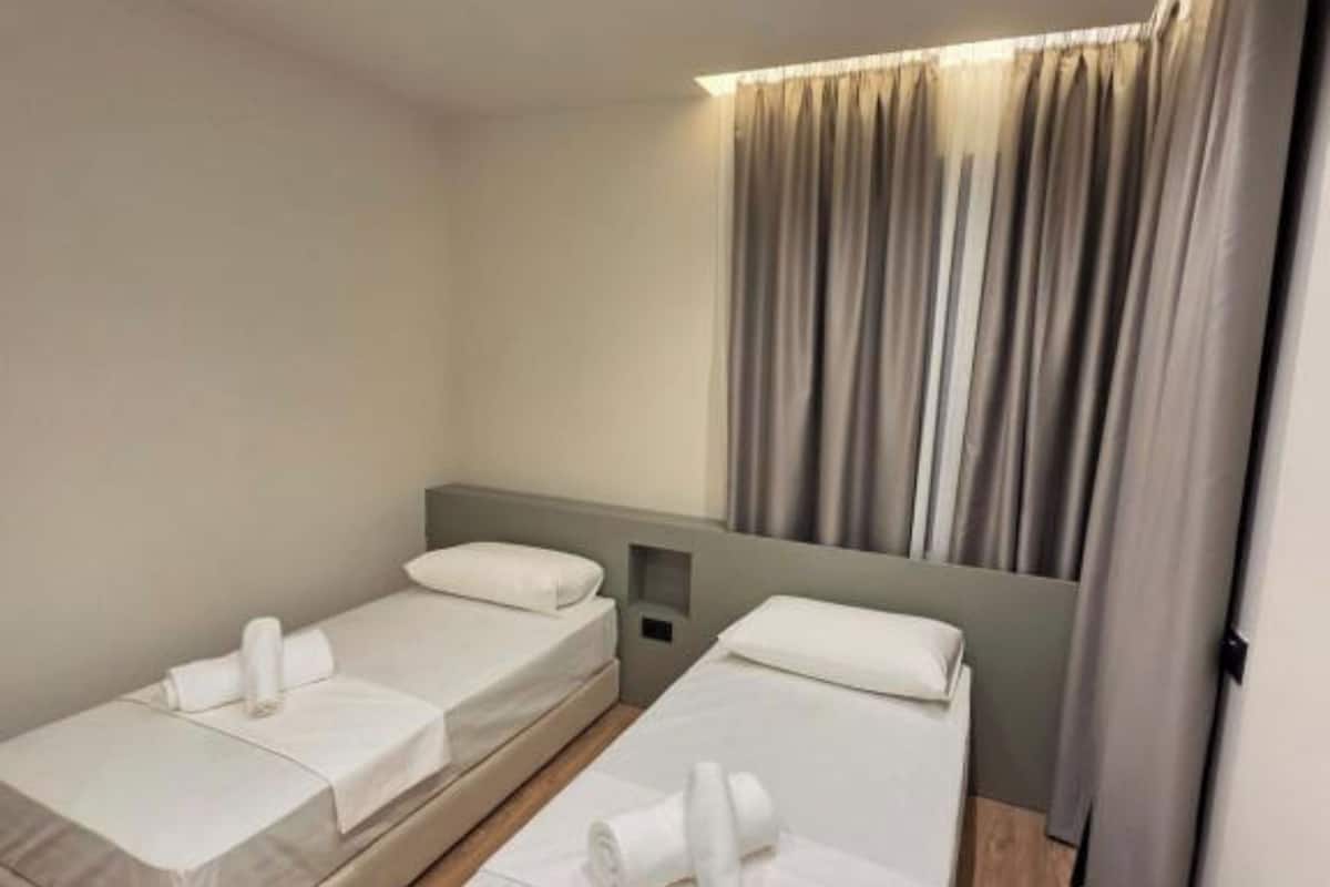 Habitación de hotel con dos camas individuales, una mesita de noche pequeña, una lámpara de pared y una ventana con cortinas.