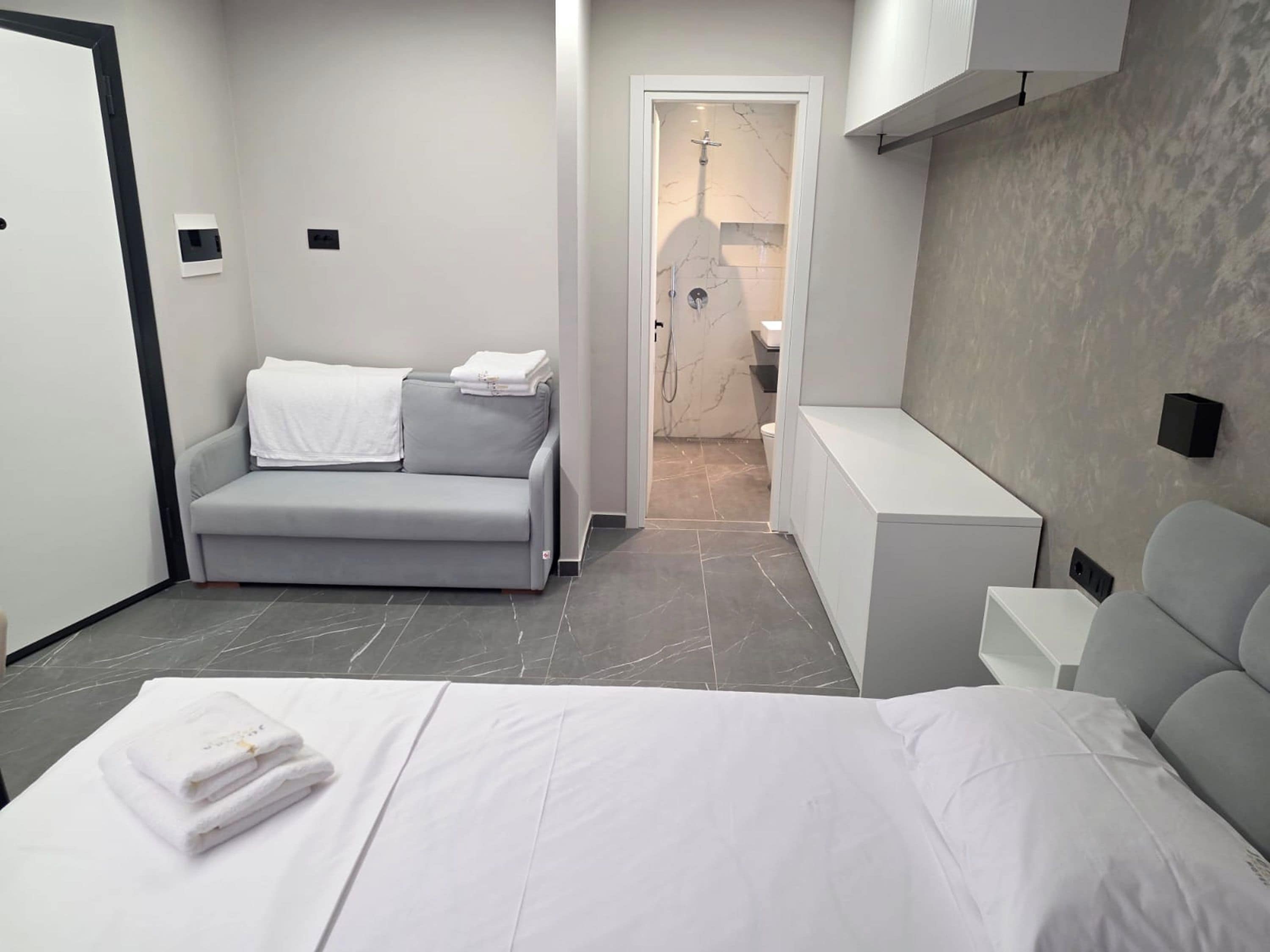 A modern hotel room with a grey sofa, a bed with white linens, a small bedside table, and a shower area.