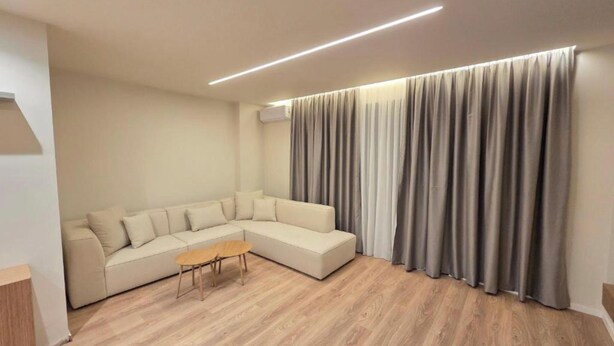 A modern living room with a sectional sofa, a small wooden table, and a large window with grey curtains. - Joanna Prestige Apartments (Sarande)