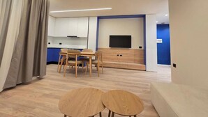 A modern living room with a sofa, two small tables, a TV, and a kitchen area with cabinets and a flat-screen TV. - Joanna Prestige Apartments (Sarande)