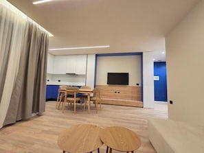 Een moderne woonkamer met een bank, twee bijzettafeltjes, een televisie, en een keukenhoek met kasten en een flatscreen-tv.