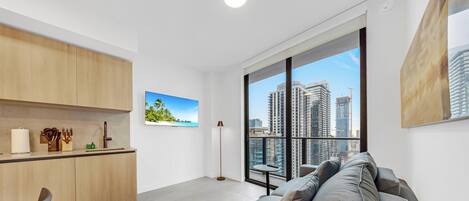 A modern living room with a large window, a kitchen area with wooden cabinets, and a city view.
