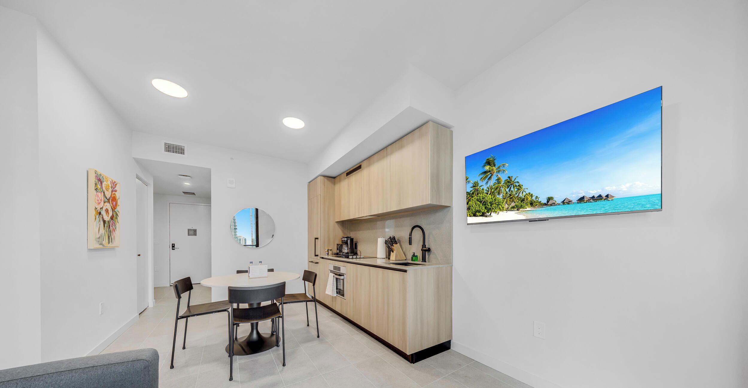 A modern kitchen with a dining area, a large window with a tropical view, and a painting on the wall.