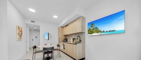 A modern kitchen with a dining area, a large window with a tropical view, and a painting on the wall.