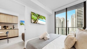 Een moderne hotelkamer met een groot bed, uitzicht op stadsgebouwen, een keukenhoek en een flatscreen televisie aan de muur.