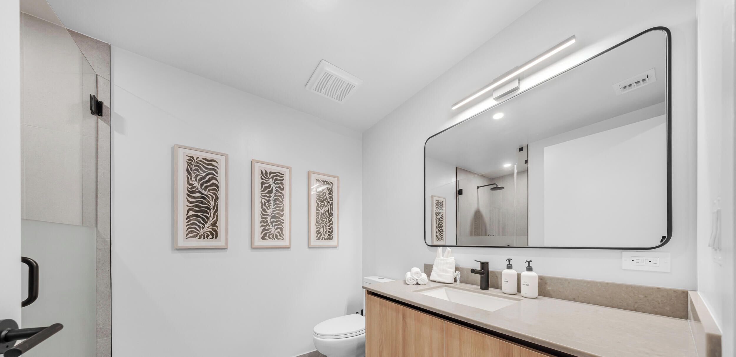 Un baño moderno con un espejo grande, tres cuadros enmarcados, un inodoro y un lavamanos con artículos de aseo.