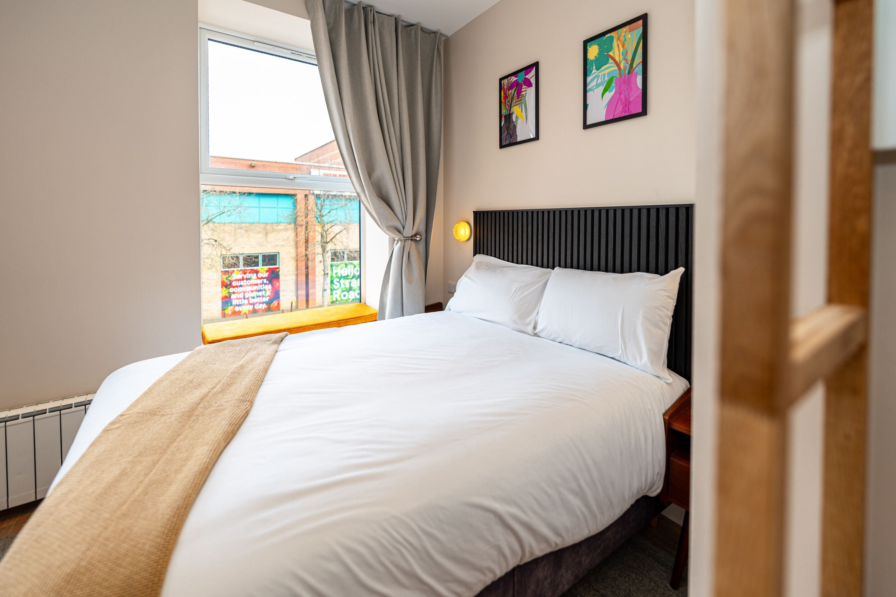 A hotel room with a bed, a window with curtains, a bedside table, and two wall-mounted artworks.