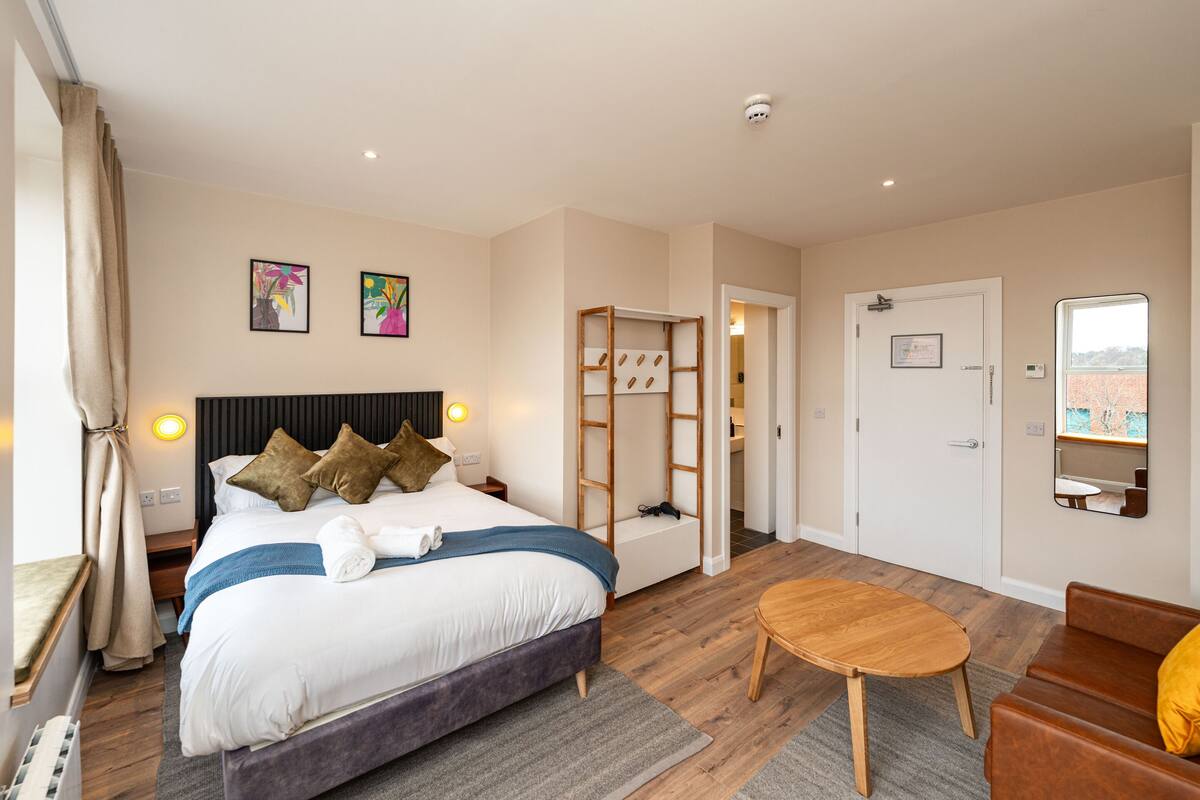 A hotel room with a bed, bedside table, lamp, and a view of the city through a window.